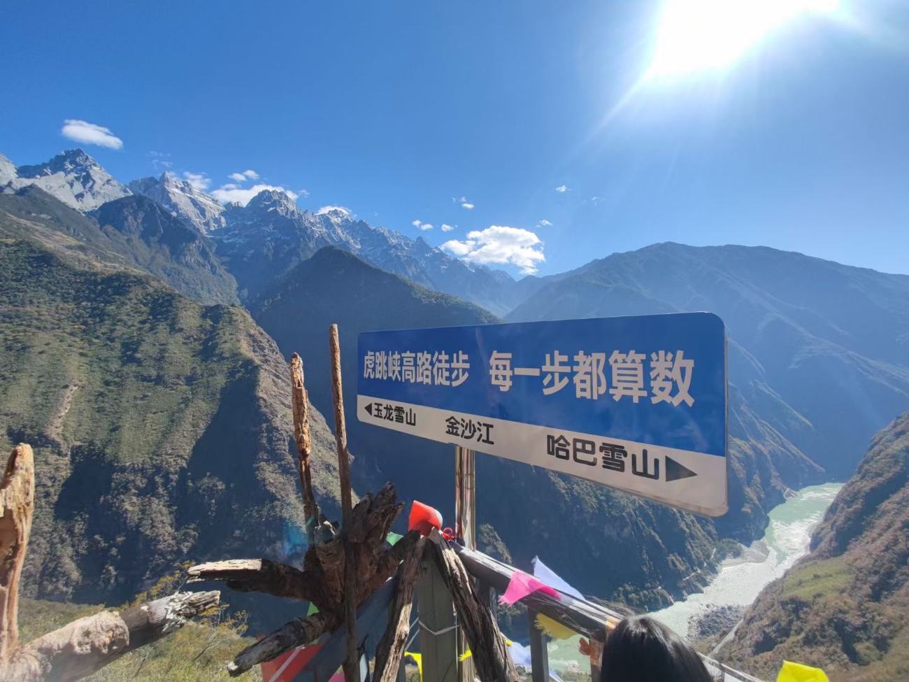 Blue trail sign reading Tiger Leaping Gorge high trail, every step counts, with arrows to Jade Dragon Snow Mountain and Haba Snow Mountain, and mountains and river behind