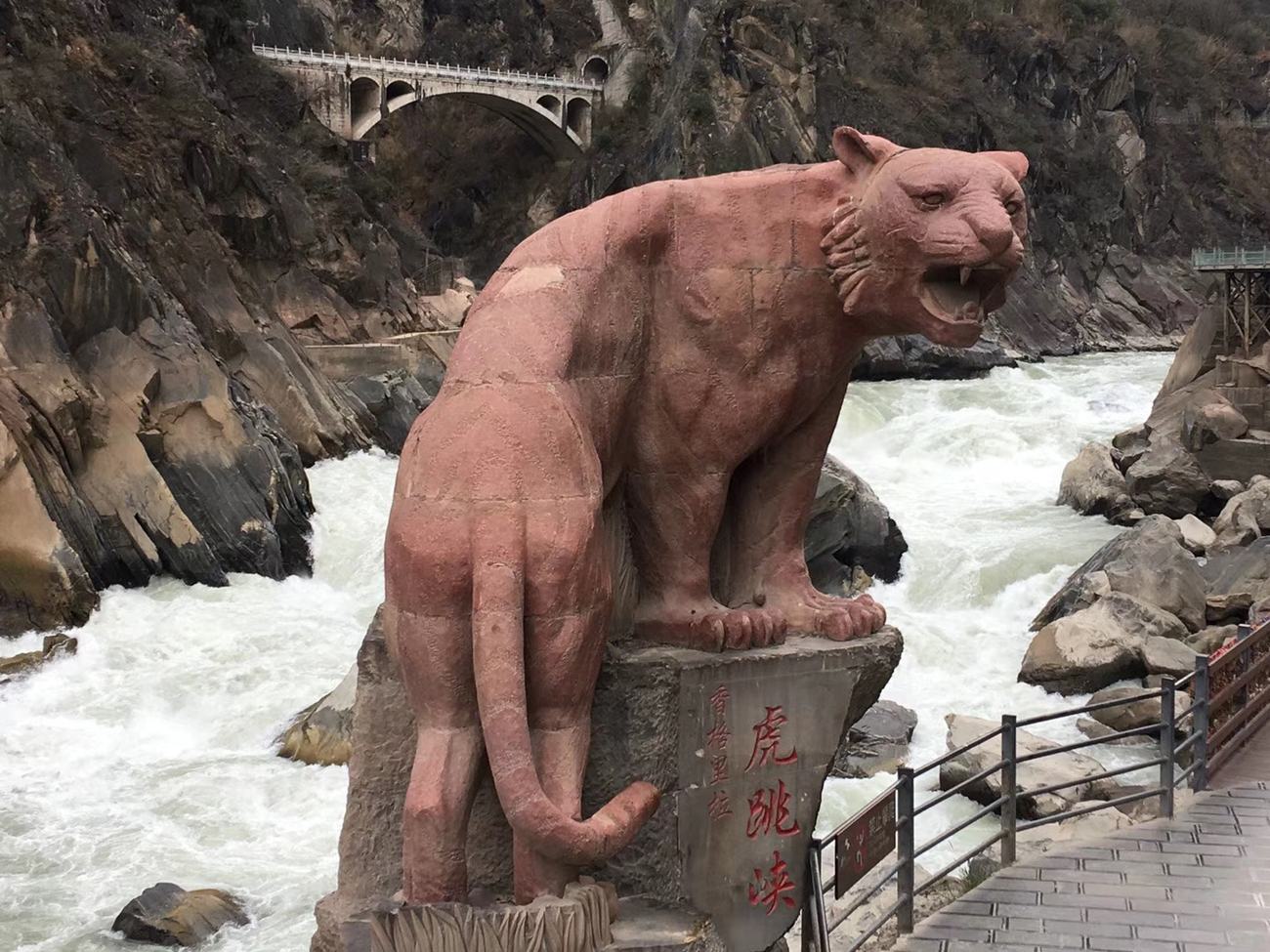 Close-up of the red stone tiger statue snarling above white rapids, with an old stone arch bridge behind